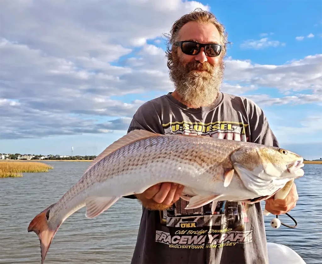 an older angler holding a large fish in front of marsh grasses and calm water