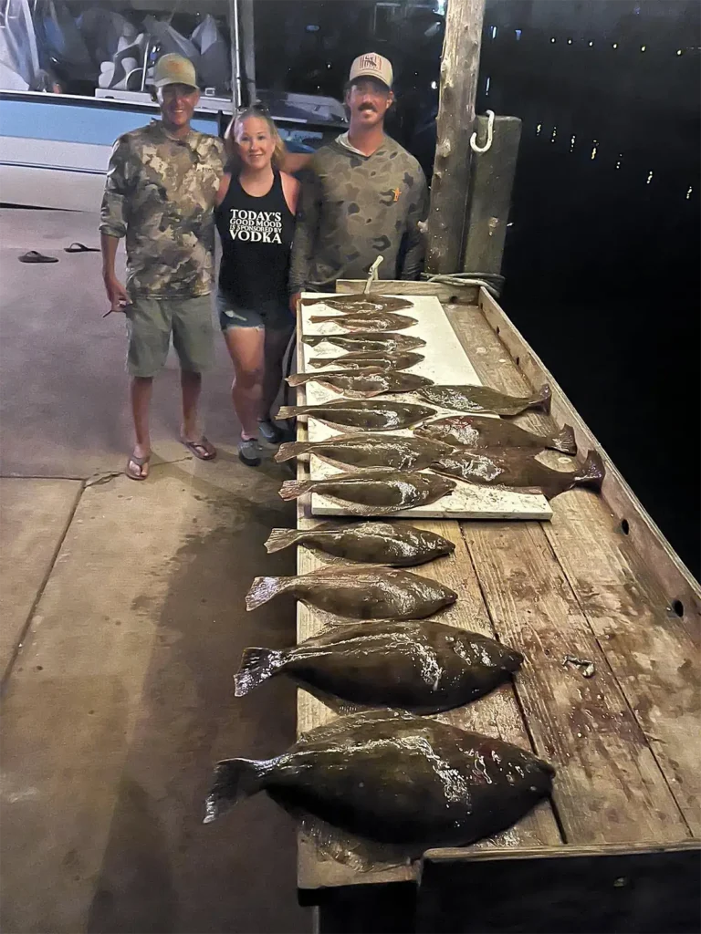two men and one woman standing beside a dockside fish cleaning table with multiple flounder laid out in a row at night