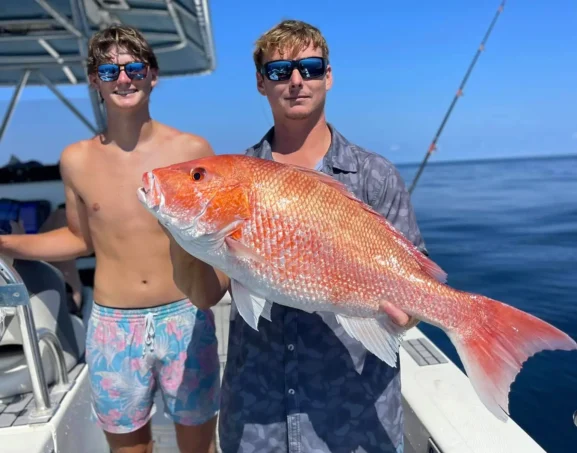 man holding a red snapper on a fishing boat with another man standing nearby