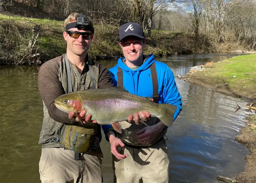 Two anglers in waders holding a large rainbow trout beside a creek