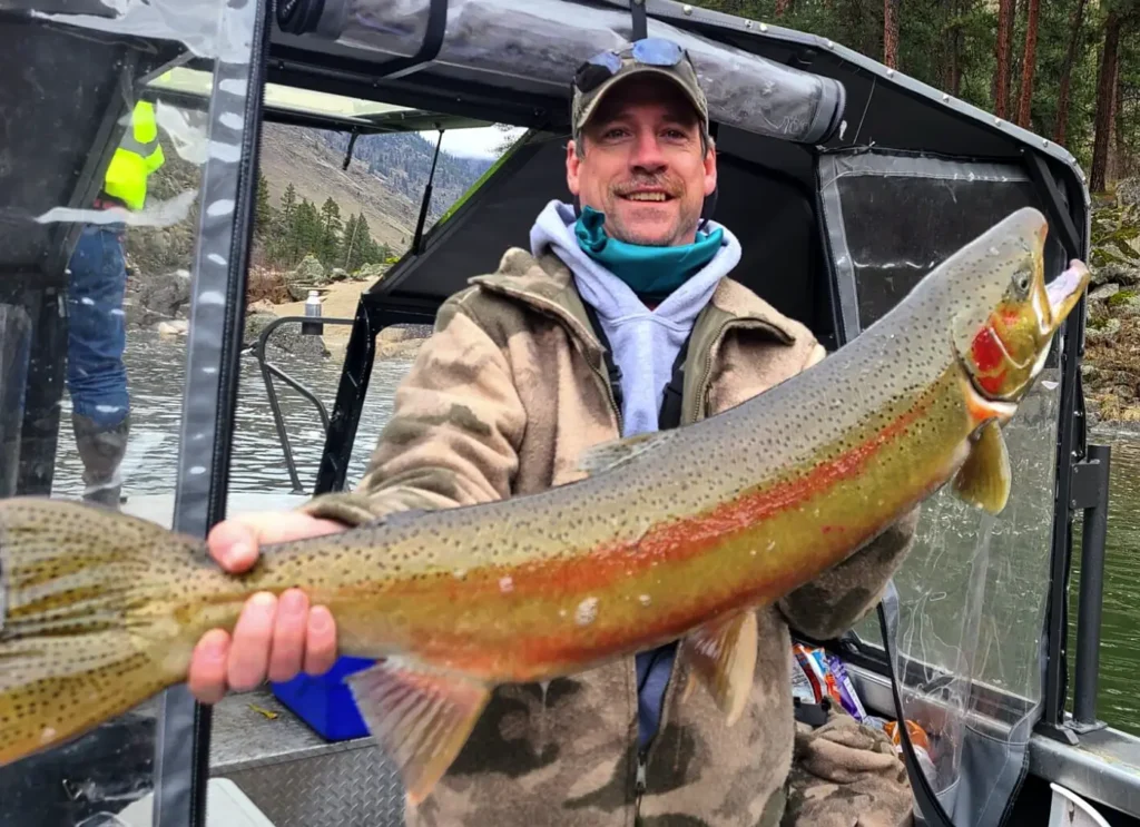 Angler holding a brightly colored Idaho steelhead beside a jet boat in cold weather