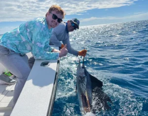 two anglers holding a sailfish beside a boat on clear offshore water