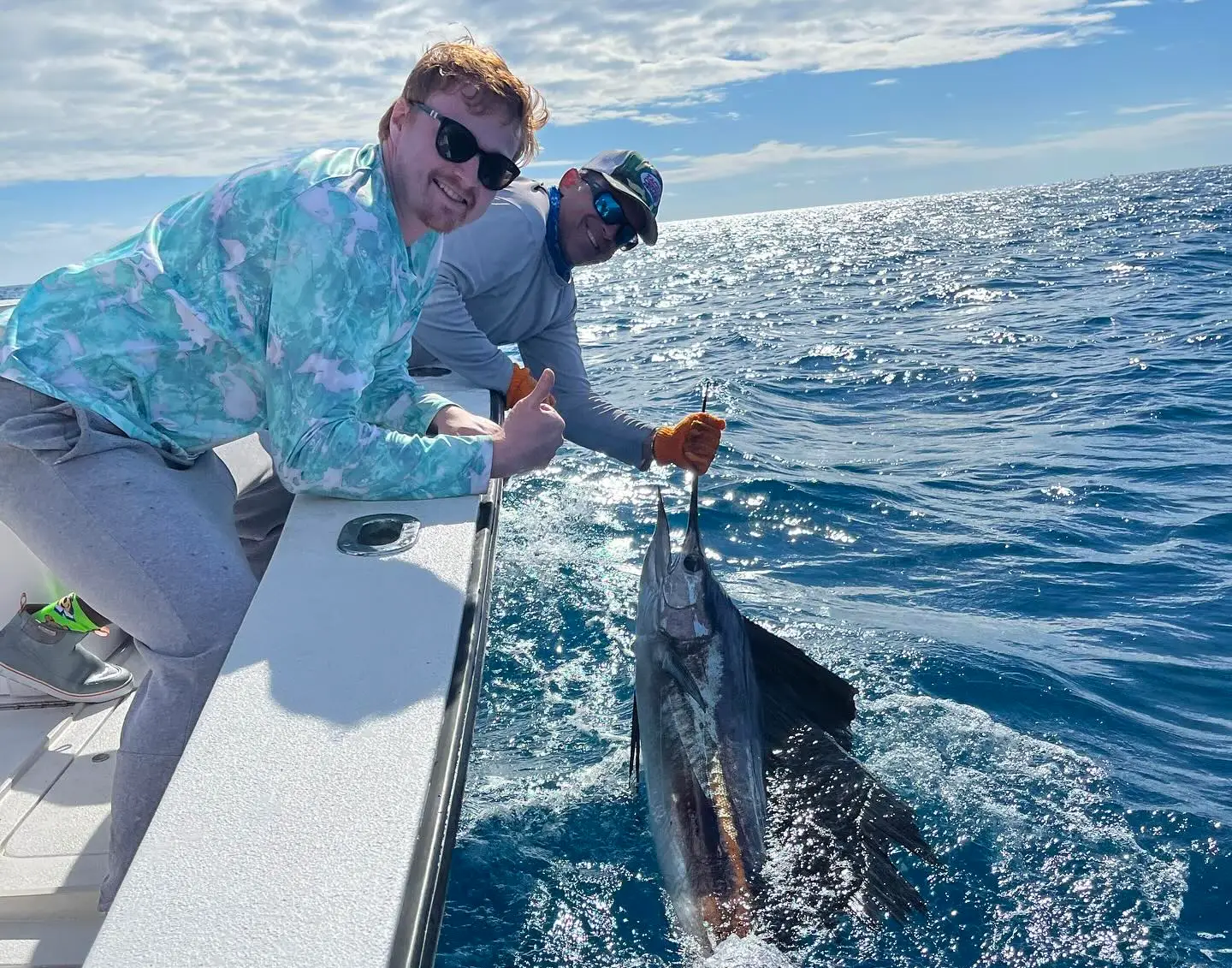 two anglers holding a sailfish beside a boat on clear offshore water