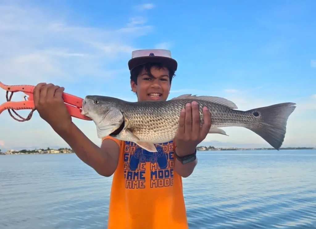 young angler holding a redfish while standing near shallow coastal water