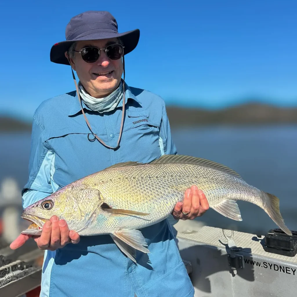 Angler wearing a hat and sunglasses holding a large fish on a boat in calm inshore water near Sydney