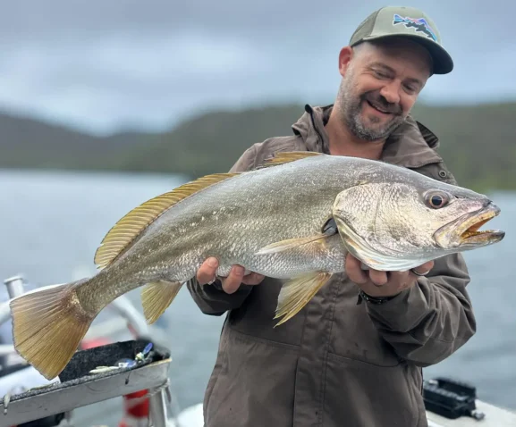 Angler holding a large silver fish on a boat with calm water and shoreline in the background near Sydney