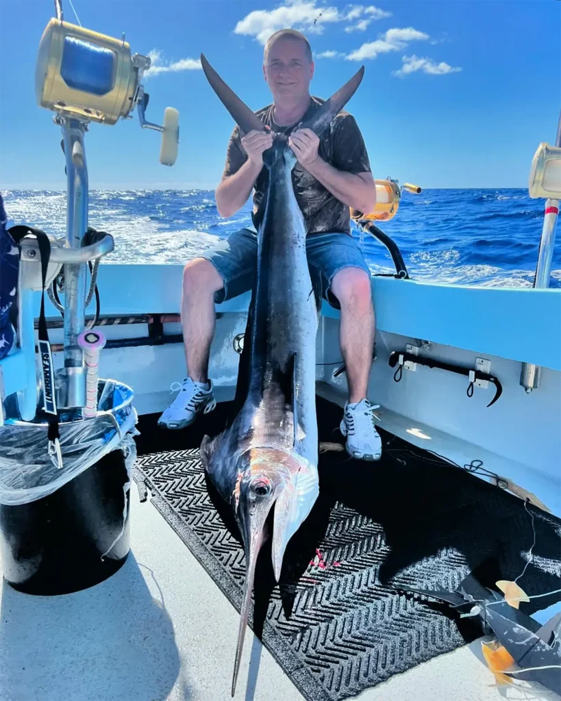 Angler seated on a fishing boat holding a large swordfish in open ocean conditions